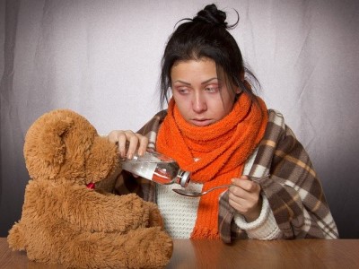 Eine junge Frau mit Erkältung nimmt Medizin und schaut auf einen Teddybären